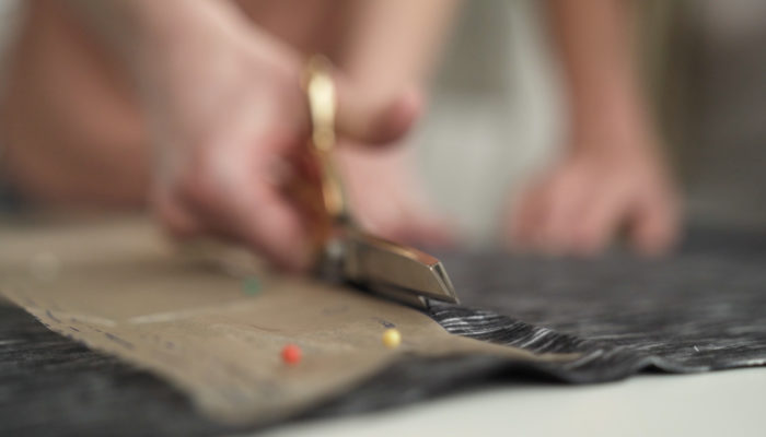 A person with fabric scissors cutting a fabric pattern out of a heathered gray cloth.