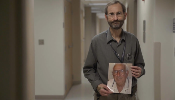 A bearded man with glasses holding a photo of an older, white haired man.